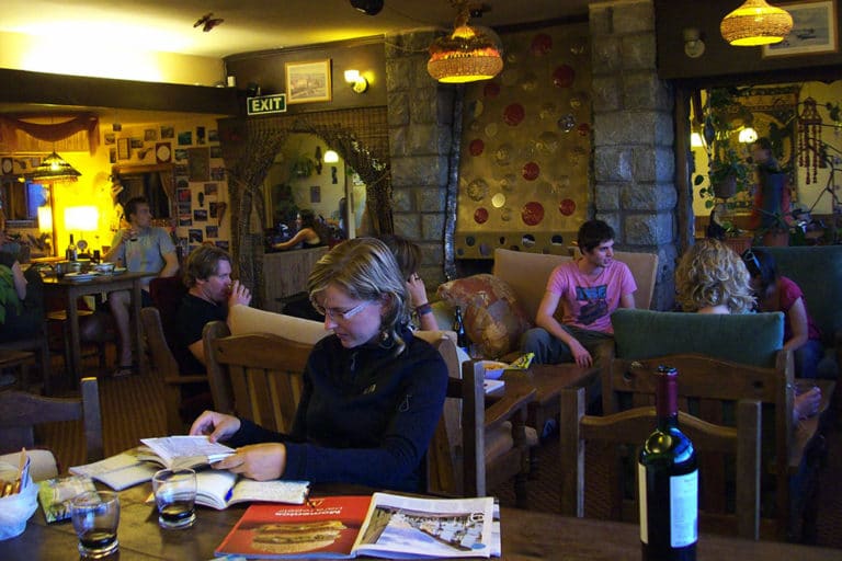 Huesped sentada a la mesa leyendo y un grupo de huespedes reunidos detras sentados en el living